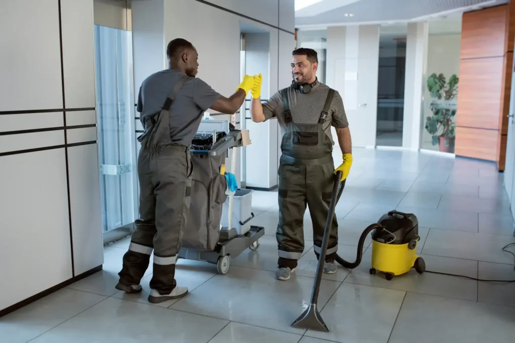 Two male commercial cleaners in gray uniforms giving a high-five in a modern office hallway next to a cleaning cart and vacuum cleaner.