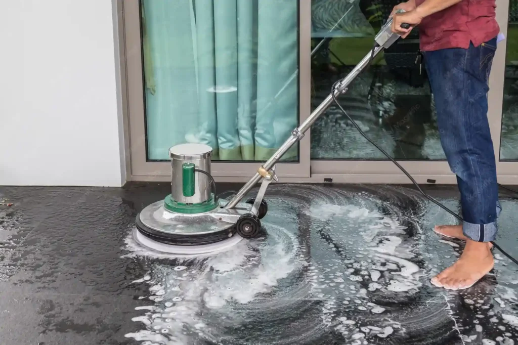 A worker operating an industrial floor buffer and polishing machine with a soapy chemical solution on a dark stone patio floor.