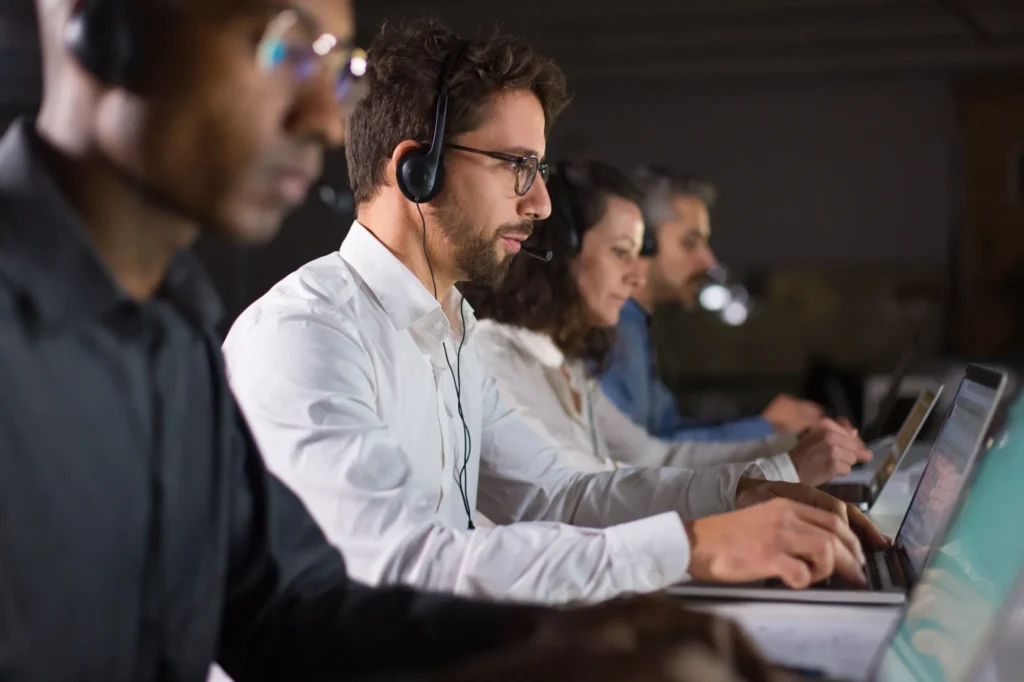 A side profile view of corporate customer support operators wearing headsets and typing on laptops in a dimly lit call center environment.