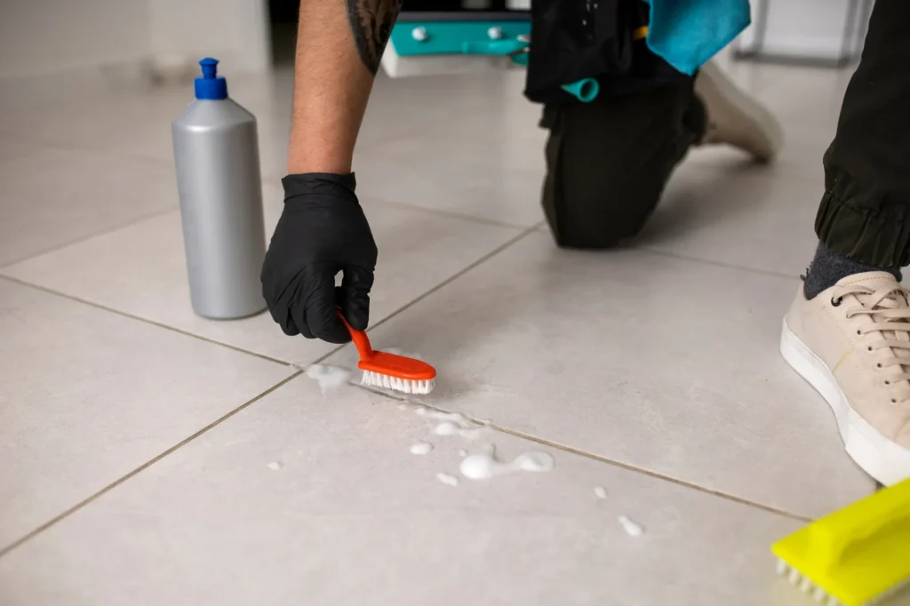 A professional cleaner wearing black gloves kneels on a tiled floor, using an orange brush and foaming solution to clean grout lines.