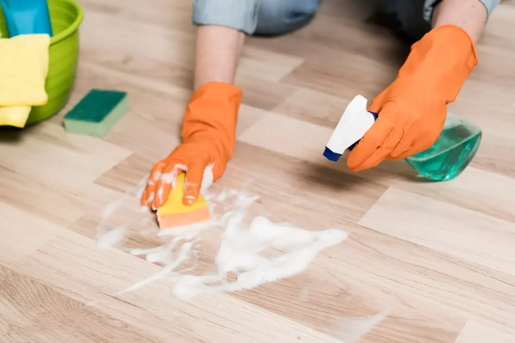 A high-angle close-up of a person wearing orange rubber gloves scrubbing a light wood laminate floor with a sponge and soapy spray solution.